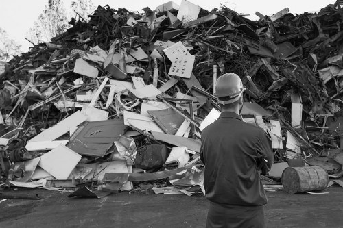 A man wearing a hard hat and dark work clothing stands in front of a large, disorganized pile of mixed waste materials on a paved surface. The debris includes various types of discarded electronic appliances, plastic items, metal scraps, and construction debris, with some items partially crushed or broken. The waste is stacked high, occupying the background and extending beyond the image frame, suggesting a significant accumulation of rubbish typical of site clearance or waste collection activities. The scene appears to be outdoors, with no visible structures or greenery in the immediate vicinity. The lighting is natural, creating contrast between the darker clothing of the individual and the lighter tones of the waste pile. This image closely relates to rubbish removal services offered by companies like House Clearance Greenhithe, serving local areas such as Greenhithe and the surrounding Kent region, emphasizing professional waste management and clearance efforts.