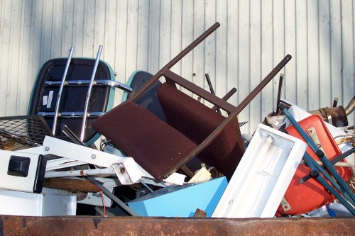 A heap of discarded household items and rubbish, including a dark brown fabric-upholstered chair with wooden legs, a white plastic window frame, a blue plastic container, and various pieces of metal and plastic debris, all piled outdoors on a weathered surface near a light-colored corrugated metal fence. The objects appear to be part of a house clearance, with some items leaning or stacked irregularly, indicative of rubbish removal activities in a residential setting. The scene is lit with natural daylight, highlighting the different textures of the materials, from the smooth plastic to the fabric and metal surfaces. This collection of waste items resembles typical household clearance rubbish that a specialist like House Clearance Greenhithe may help to remove from properties in the Greenhithe area, close to Dartford and the surrounding region.