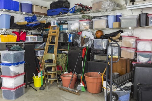 The image shows a densely packed storage area or garage filled with various household rubbish and clutter, including multiple plastic storage containers in different sizes and colours, some with lids and others open, stacked on shelves and on the floor. A small wooden stepladder leans against the shelving unit, alongside a folding chair. Several household cleaning tools such as mops, brooms, and a dustpan are visible, some standing upright and others resting against objects. There are also a few larger items, including a black plastic storage bin, a small suitcase, and miscellaneous items that appear to be discarded or waiting to be disposed of. The environment appears well-lit, with natural or artificial light illuminating the cluttered space. This scene is representative of a typical residential garage in Greenhithe where a rubbish removal service, like those offered by House Clearance Greenhithe, might be required to clear out accumulated domestic waste and personal belongings, especially in preparation for clearance or renovation projects in the area.