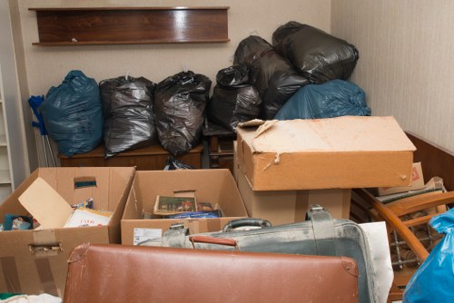 In this indoor scene, there are multiple black and blue plastic rubbish bags stacked against a light-colored wall, with some bags leaning on a small wooden cabinet. In the foreground, several open cardboard boxes contain various items, including paper documents or magazines, and a metal lamp or similar object. To the right, a partially visible wooden chair or small table appears to be used for storage or staging. The environment suggests a cluttered area likely intended for rubbish collection or house clearance purposes, with a section of a wooden shelf mounted on the wall above the bags. The lighting is neutral, highlighting the different textures of the bags, cardboard, and wood, and the overall setting indicates an active rubbish or waste removal scene typical of professional services such as those provided by House Clearance Greenhithe for local properties in Greenhithe or nearby areas.