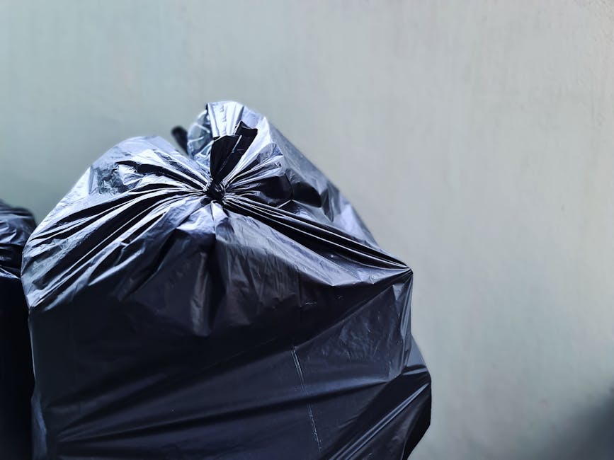A pile of yellow plastic rubbish bags filled with waste sits on a concrete sidewalk next to a weathered, graffiti-covered brick wall on the left. The bags appear to contain household waste and are tie