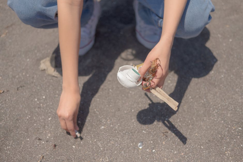 A person wearing light blue jeans and white sneakers is crouching on an outdoor paved surface, holding a small collection of rubbish in their right hand. The rubbish includes a white disposable cup wi