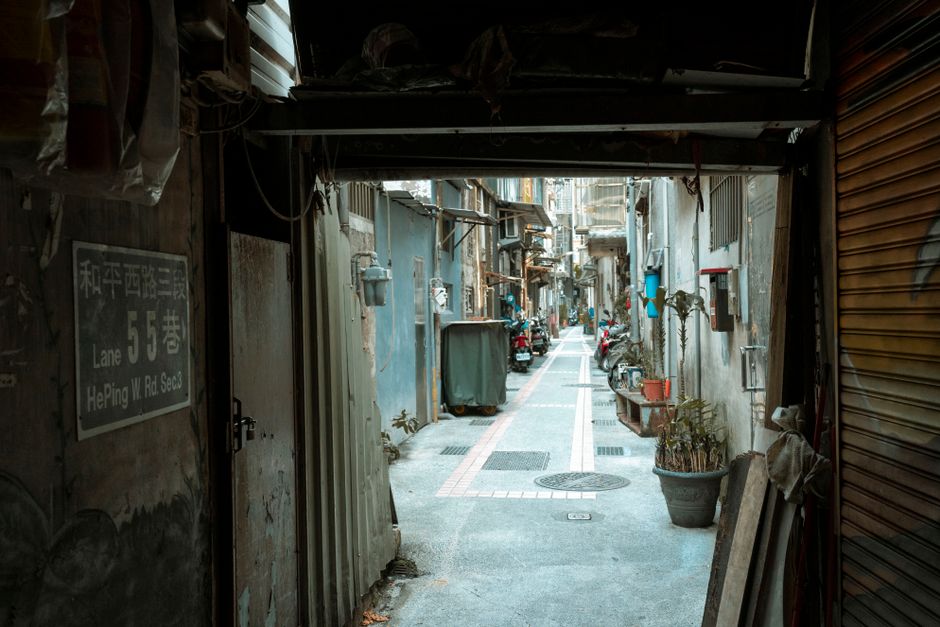 A large, worn, beige cloth bag filled with trash is placed on the pavement next to a wall in a narrow urban alleyway. The bag appears to be made of thick fabric, heavily stained and creased, with some