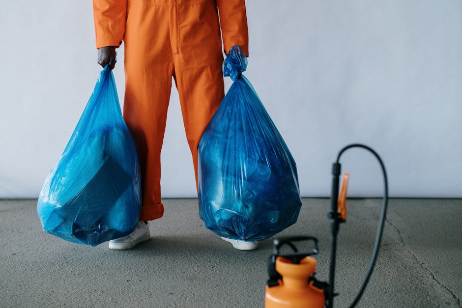 Close-up view of a person wearing orange protective overalls and white gloves, holding a large blue plastic rubbish bag with their right hand, which is partially covered by a crumpled blue plastic glo